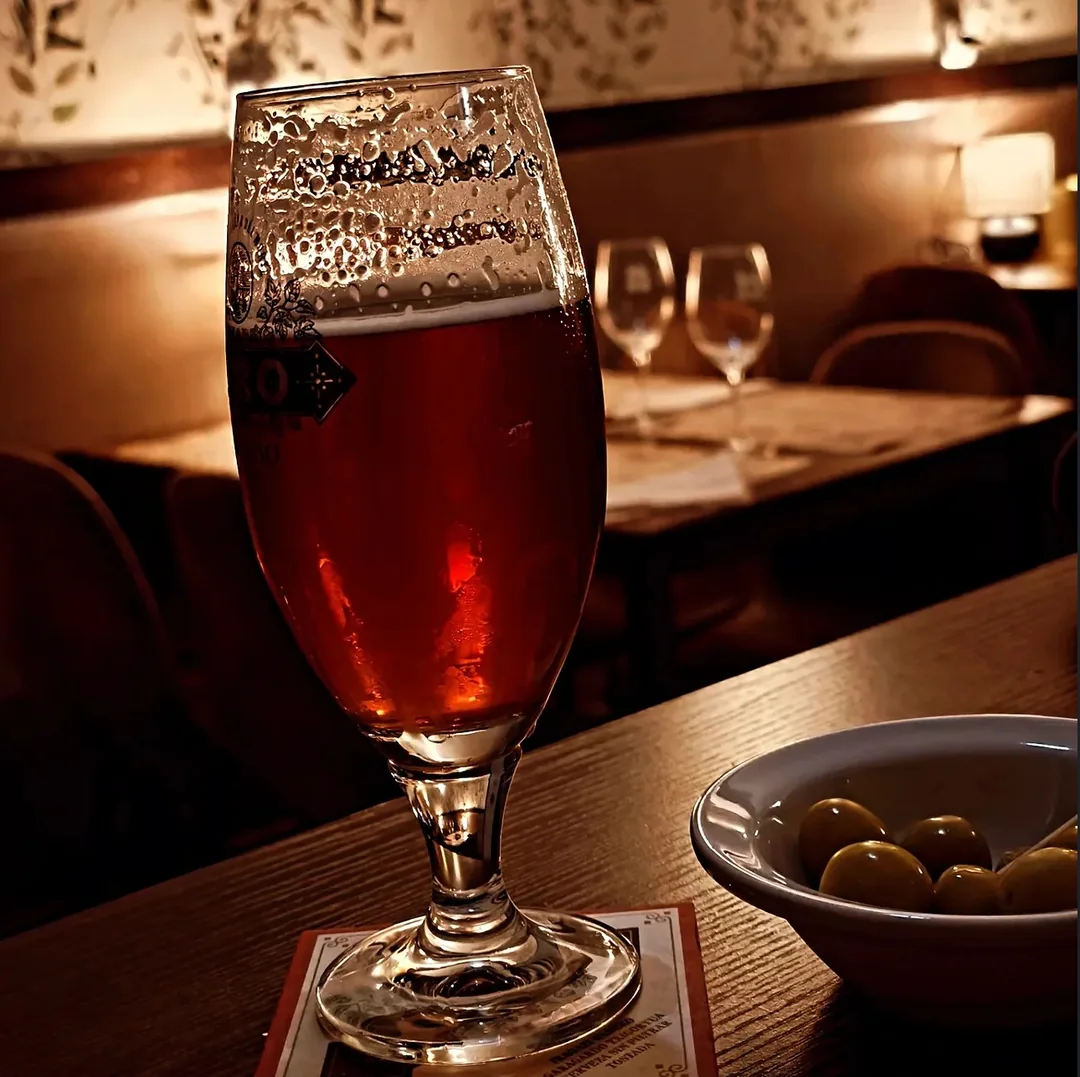 Una copa de cerveza y unas aceitunas, con luz cálida al fondo.